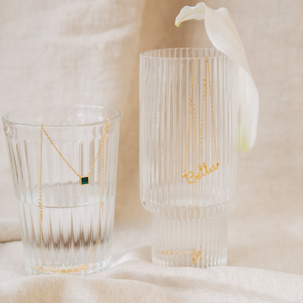 A gold Malachite Necklace with a vibrant green gemstone and a Bella nameplate necklace drape over clear glasses, capturing the allure of gemstone jewelry.