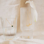 A gold Malachite Necklace with a vibrant green gemstone and a Bella nameplate necklace drape over clear glasses, capturing the allure of gemstone jewelry.