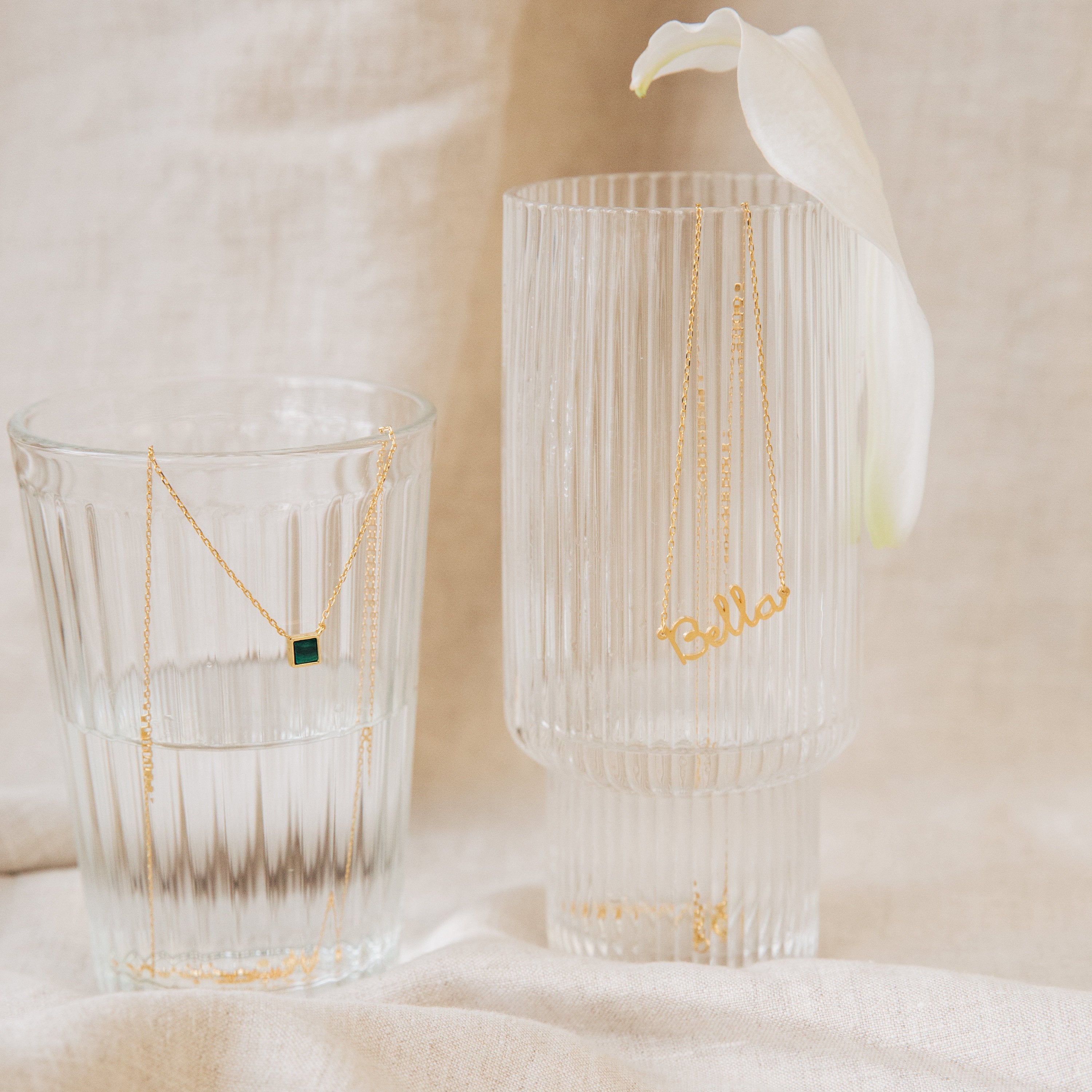 A gold Malachite Necklace with a vibrant green gemstone and a Bella nameplate necklace drape over clear glasses, capturing the allure of gemstone jewelry.