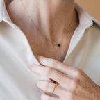 A person in a white shirt touches a Malachite Necklace with a green pendant and a matching gold gemstone ring.
