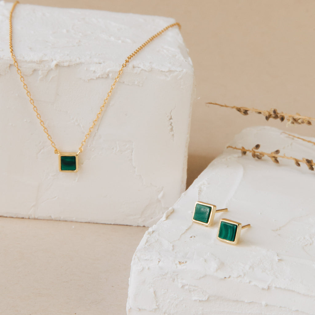 The Malachite Necklace features green square stones set in 925 sterling silver, displayed on white textured blocks—an elegant example of gemstone jewelry.
