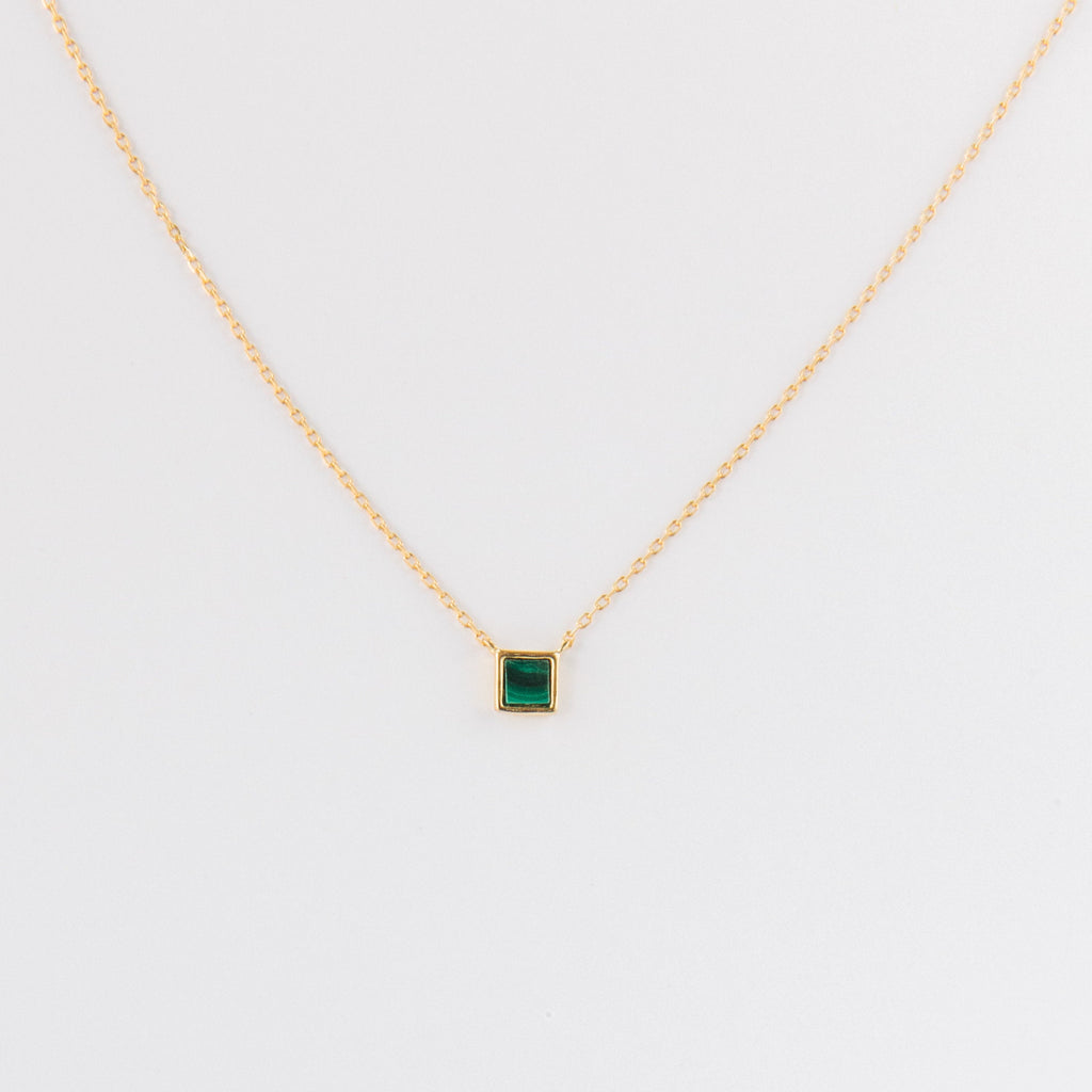 The Malachite Necklace features a gold chain and a small square malachite crystal pendant, shown on a plain white background.