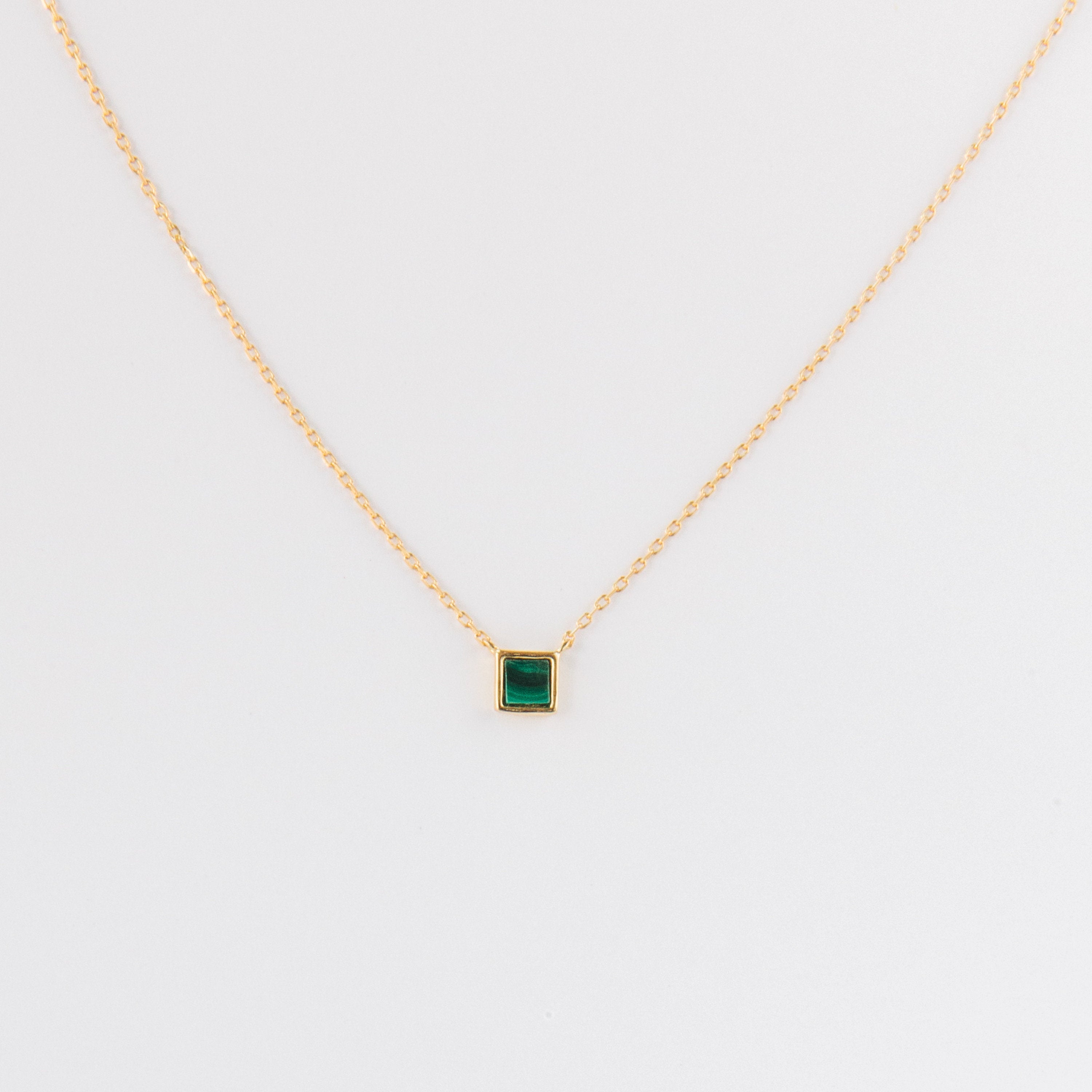 The Malachite Necklace features a gold chain and a small square malachite crystal pendant, shown on a plain white background.