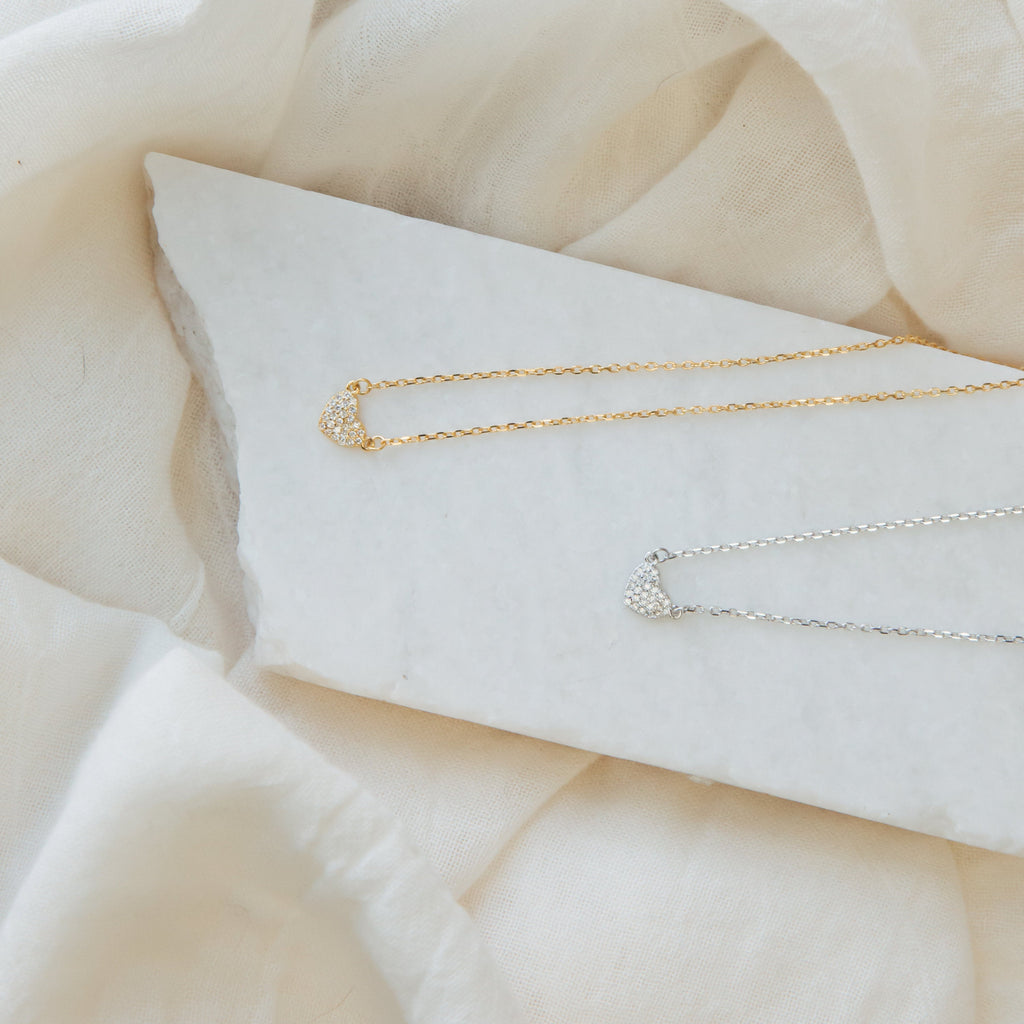 The Pave Heart Necklace, shown in gold and silver, rests on a white stone with a soft beige fabric background, highlighting its delicate and minimalist heart pendant design.