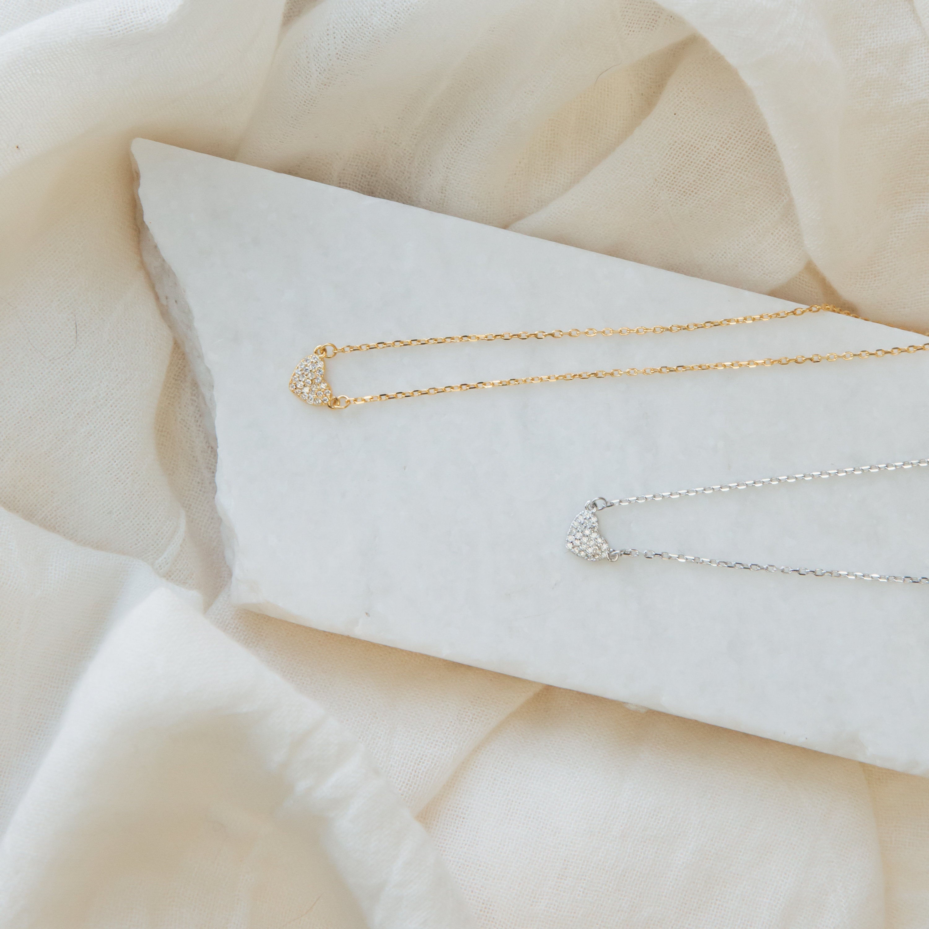 The Pave Heart Necklace, shown in gold and silver, rests on a white stone with a soft beige fabric background, highlighting its delicate and minimalist heart pendant design.