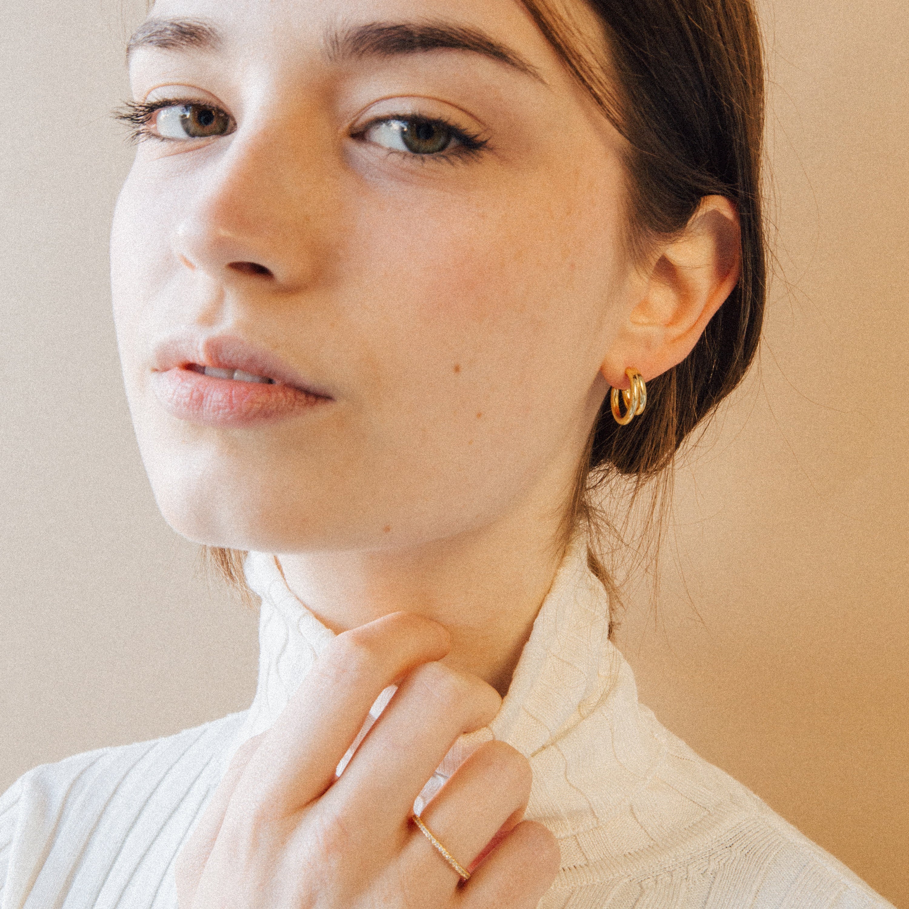 A woman with light skin wears a white turtleneck, gold ring, and Zoey Duo Hoops for a chic stacked ear look, gazing at the camera.