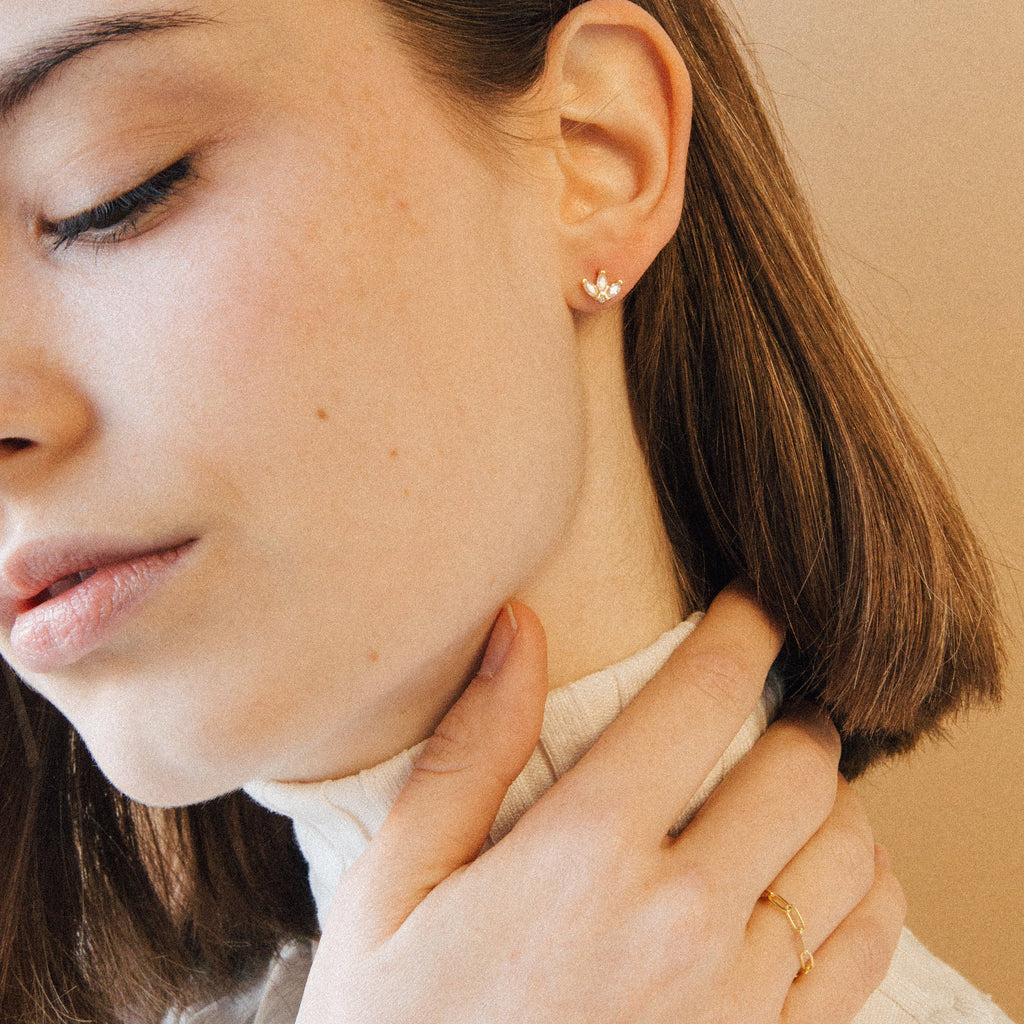 A woman with light skin wears a white turtleneck, Arya Petal Studs earrings, and a thin gold ring as she touches her neck.