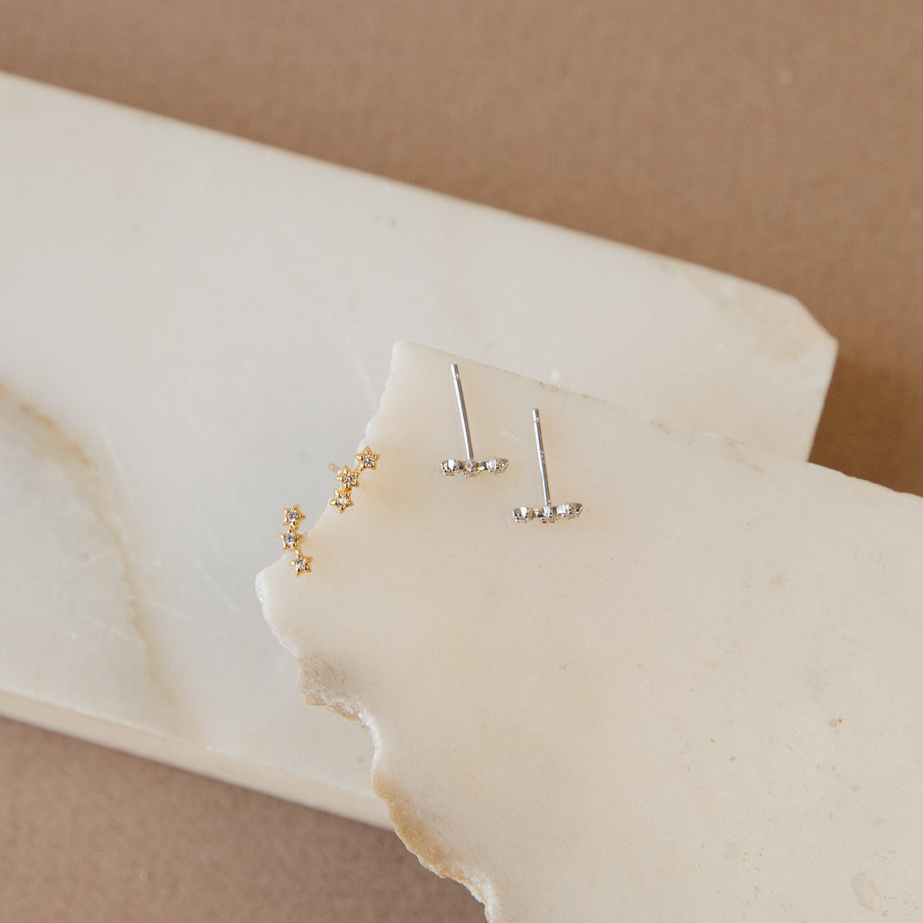 Two pairs of dainty Stella Star Studs, in gold and silver, are elegantly displayed on stacked white marble slabs.