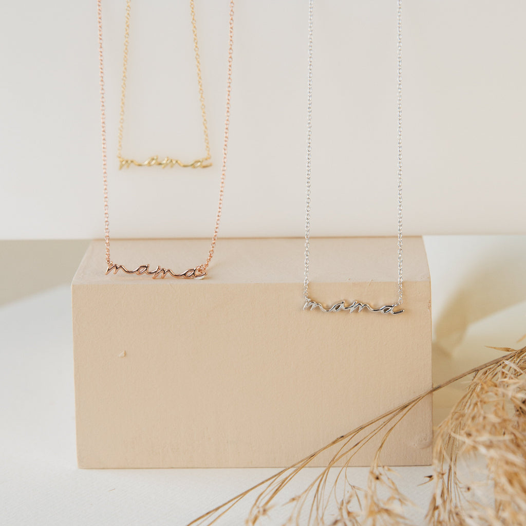 The Dainty Mama Necklace in 18K Gold, featuring a custom script font, is displayed on a beige block with dried grass in the foreground.