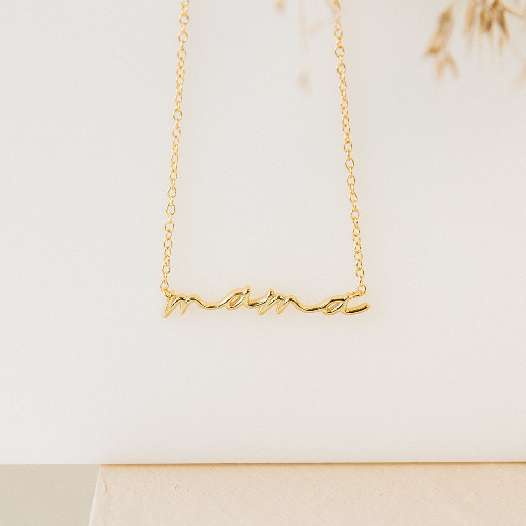 Gold necklace featuring the word “mama” in elegant cursive script, suspended from a delicate chain. Displayed against a clean, neutral background with soft lighting.