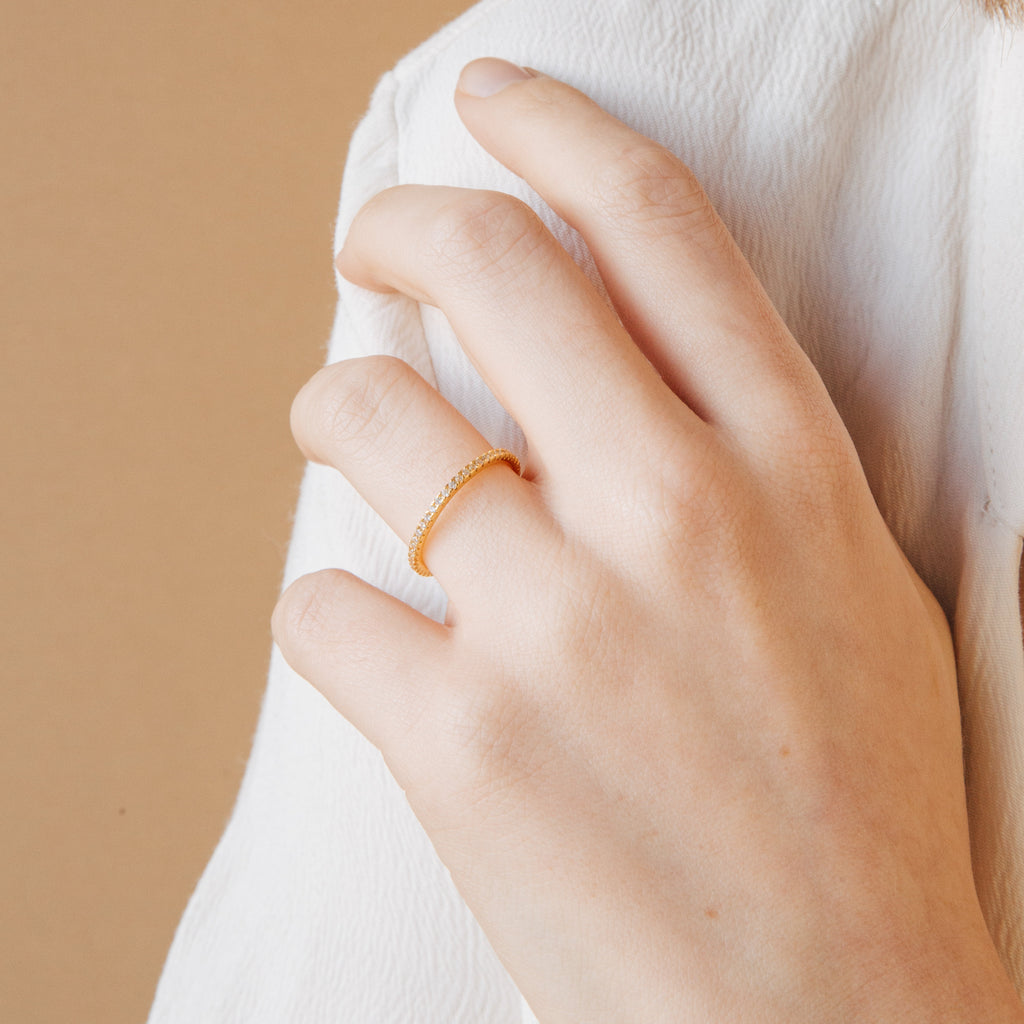 A hand wearing the Dove Eternity Ring, a petite gold band on the ring finger, set against a beige background.