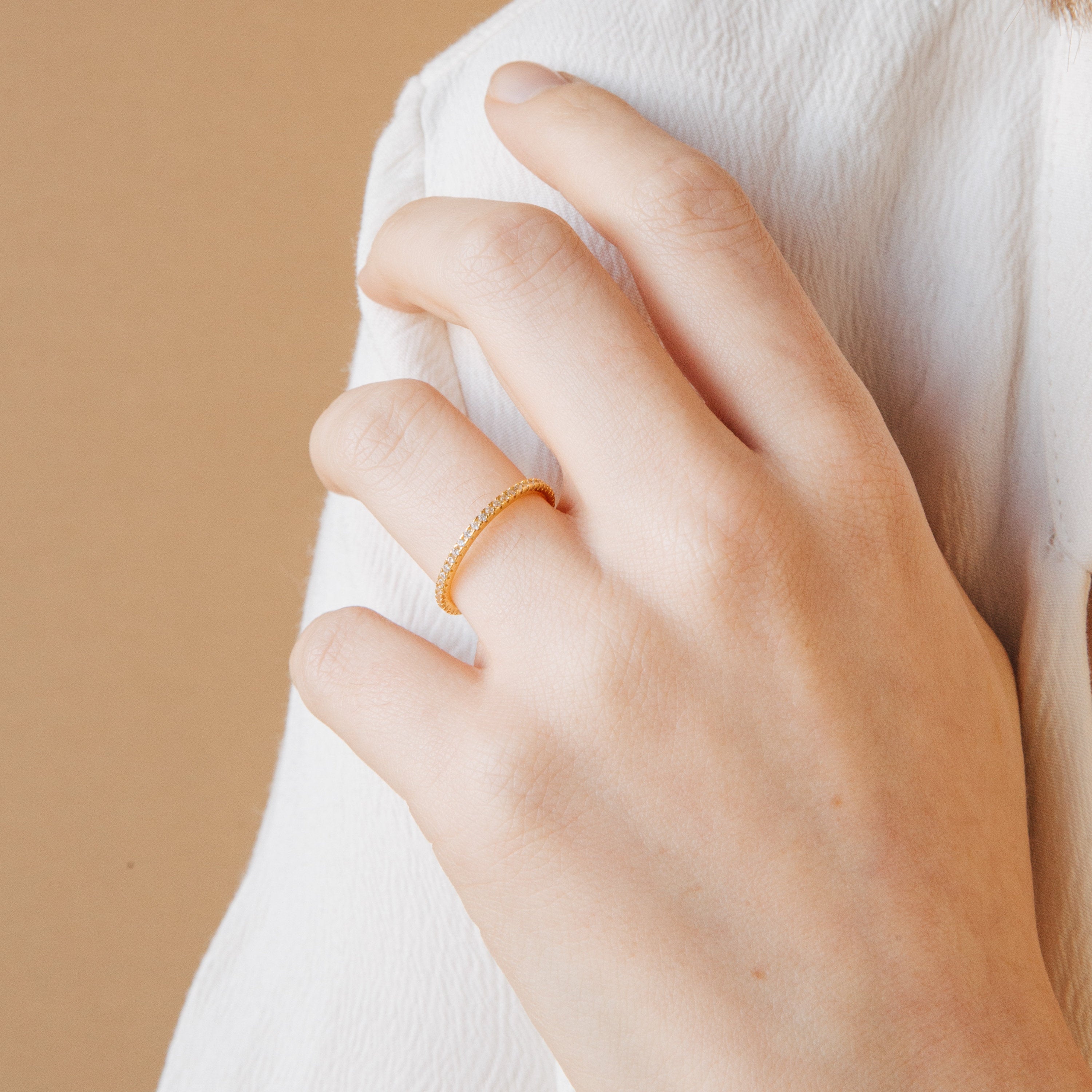 A hand wearing the Dove Eternity Ring, a petite gold band on the ring finger, set against a beige background.