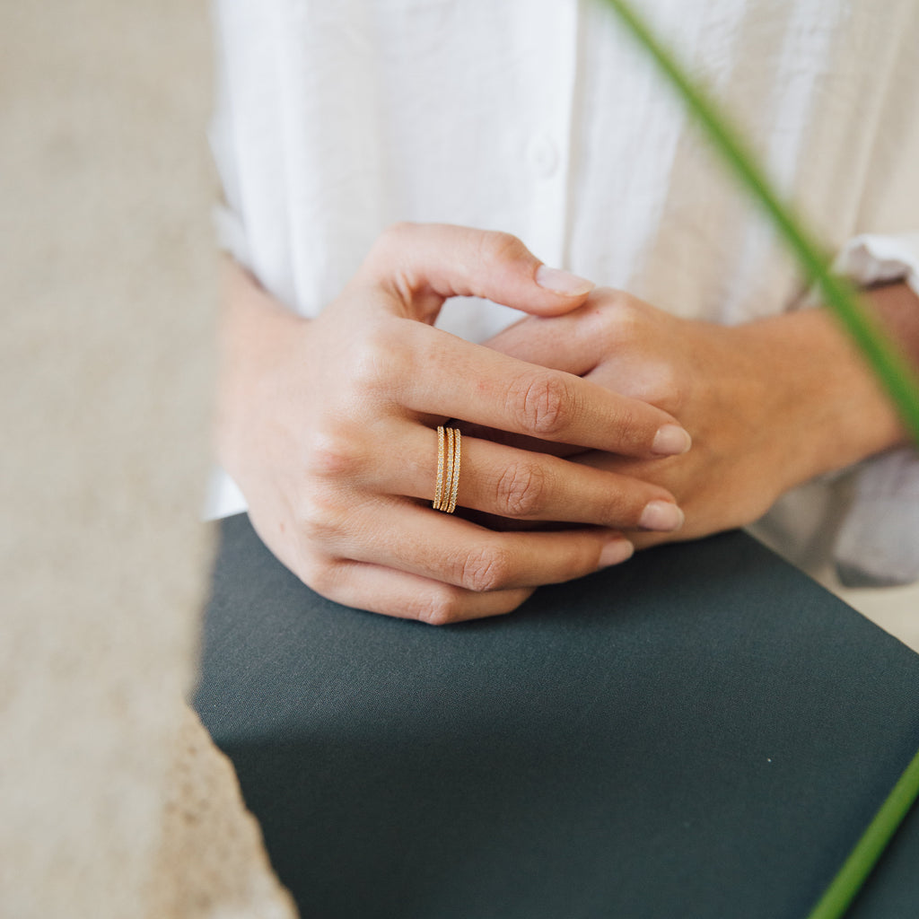 A person wearing a white shirt rests their hands, adorned with a Dove Eternity Ring, on a dark surface; a blurred plant appears in the foreground.