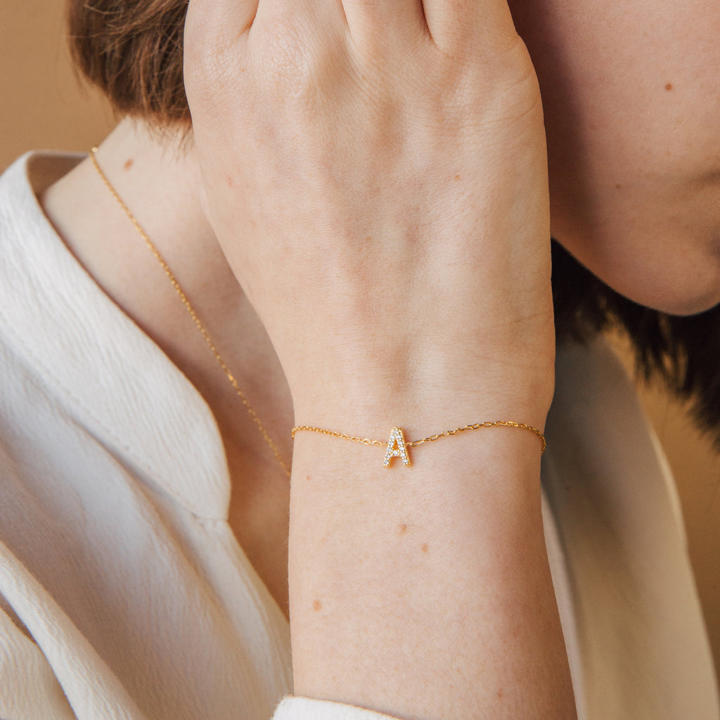 A woman wears the Pave Initial Bracelet with a letter A charm and a matching gold necklace, touching her ear.