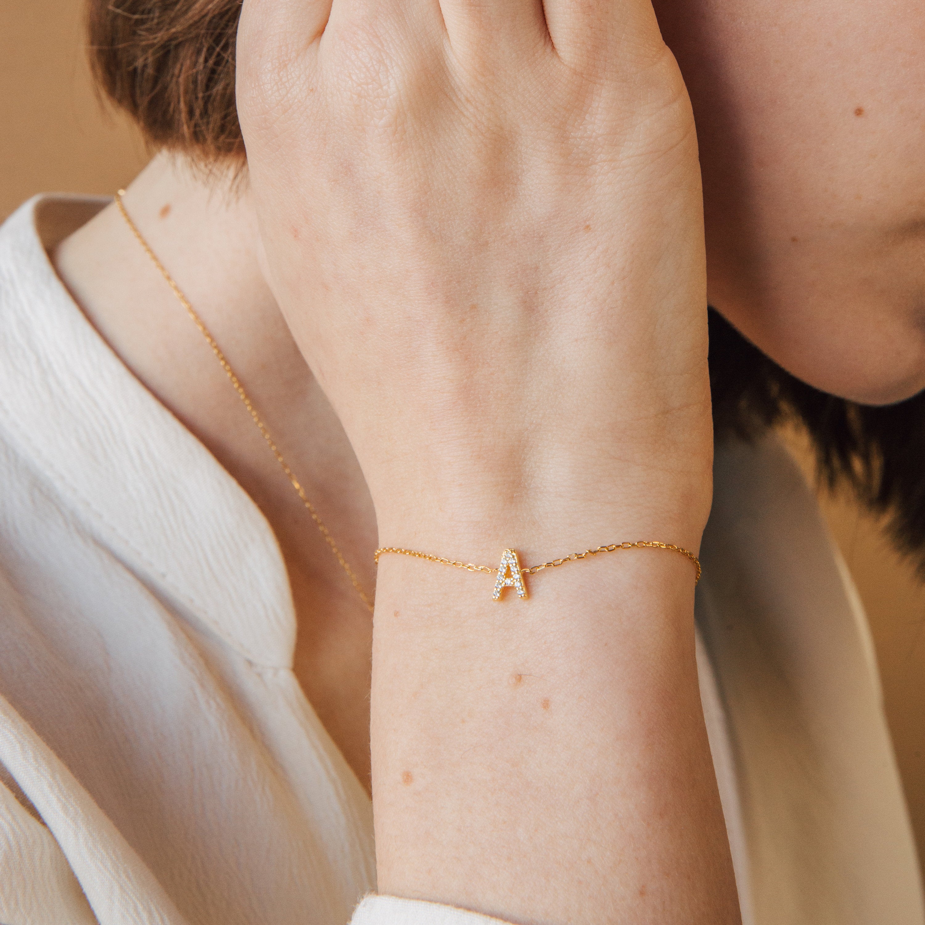 A woman wears the Pave Initial Bracelet with a letter A charm and a matching gold necklace, touching her ear.