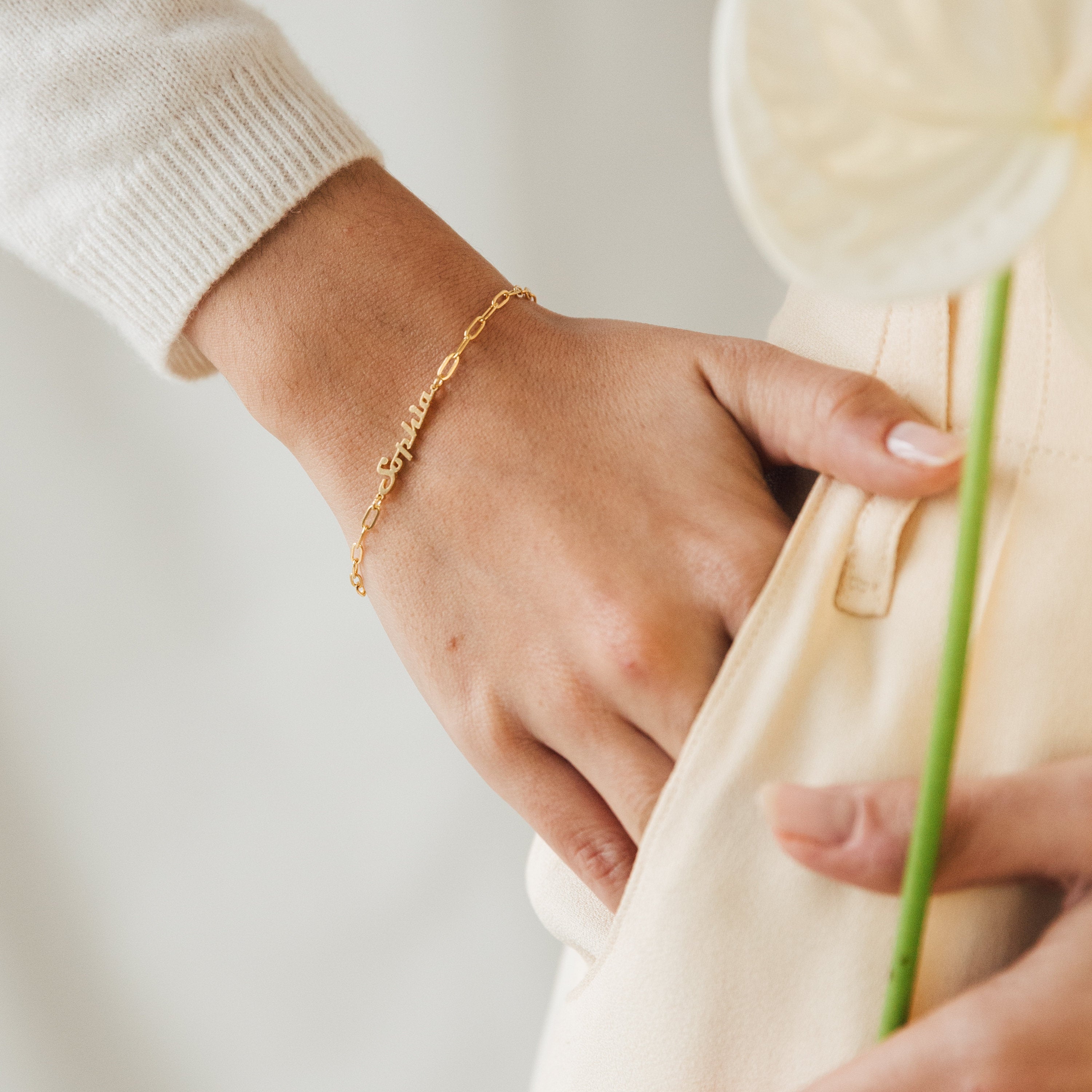 A hand adorned with the Name Link Bracelet in gold rests on a cream pocket, gently holding a pale flower stem—showcasing this modern piece of jewelry.