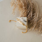 The Ladybirds Name Curb Bracelet in gold and a silver curb chain bracelet are displayed on a beige block with dried grass, highlighting their modern style.