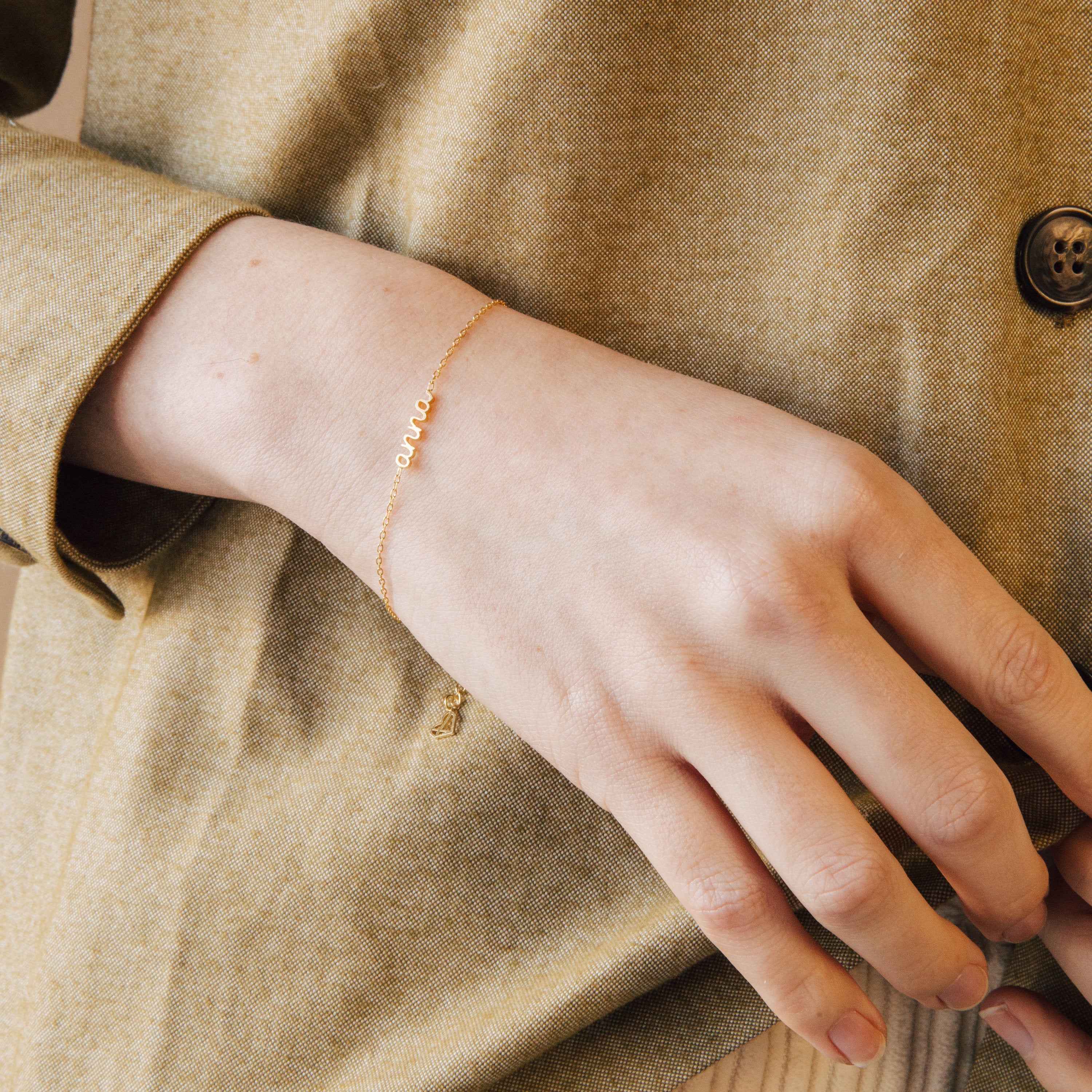 A hand wears the Petite Tiny Name Bracelet, featuring small letters, resting on a beige fabric jacket—a perfect minimal jewelry choice.