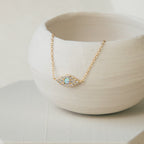 Gold necklace with an opal center set in an eye shaped pendant, bordered by a row of small clear crystals, displayed draped over a matte white ceramic bowl.