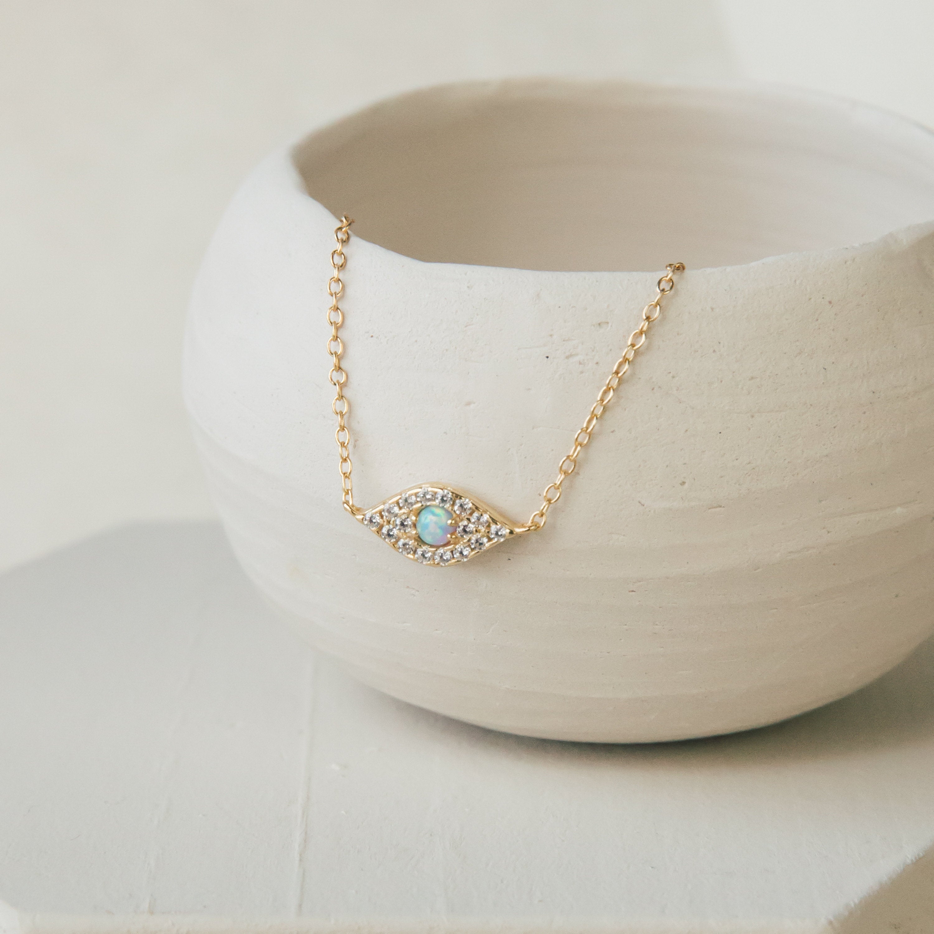 Gold necklace with an opal center set in an eye shaped pendant, bordered by a row of small clear crystals, displayed draped over a matte white ceramic bowl.