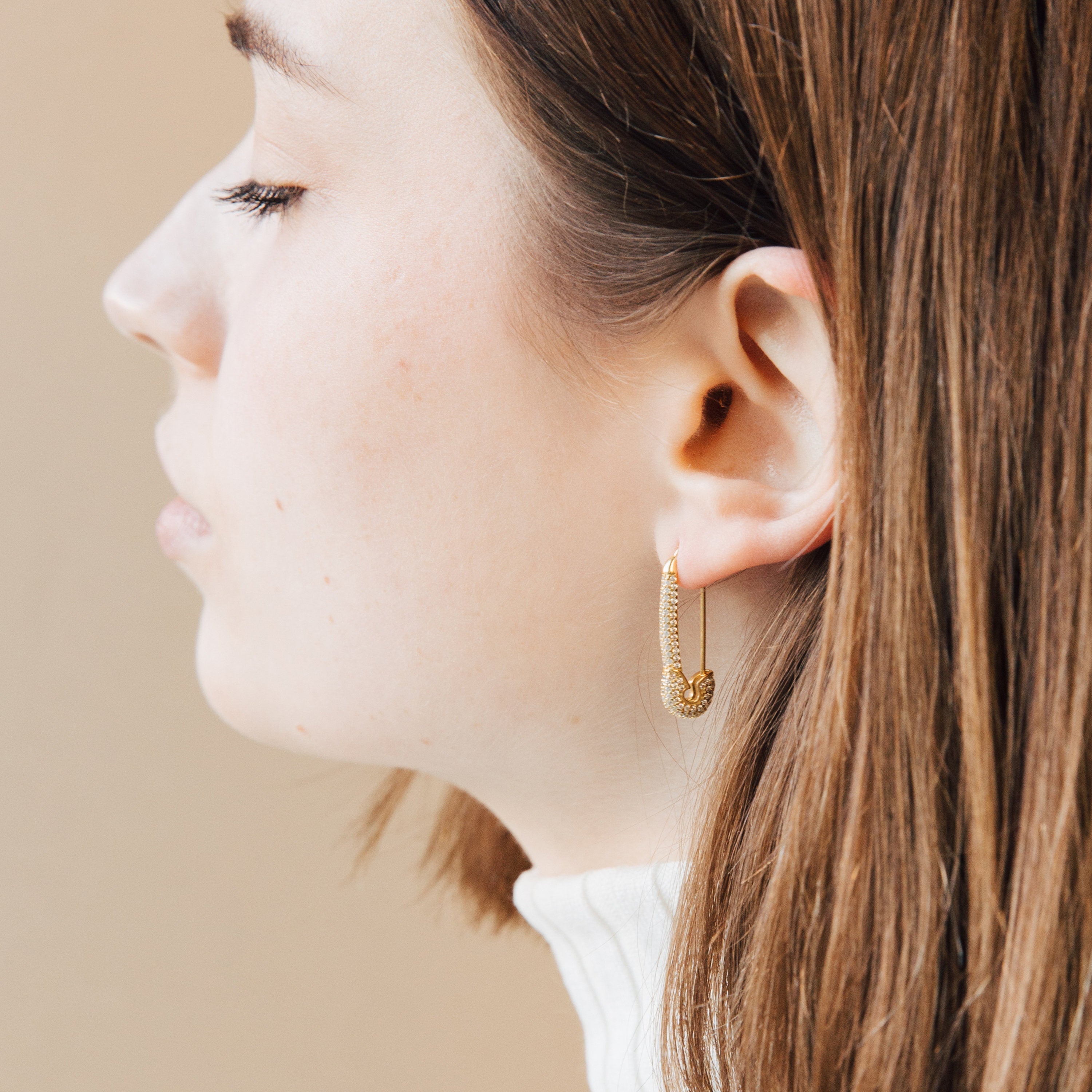A woman in profile wears Luxx Earrings—a gold safety pin design—with straight brown hair and a cream turtleneck sweater.