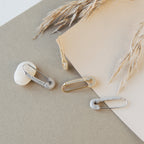 The Safety Pin Earrings Set is displayed on neutral paper with a white pebble and dried wheat stems, showcasing their minimalist charm.