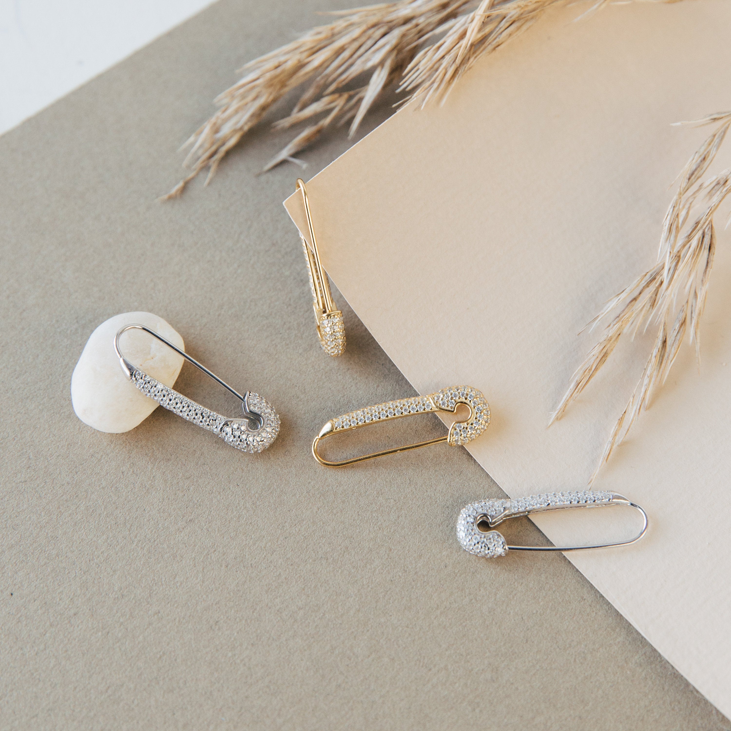 The Safety Pin Earrings Set is displayed on neutral paper with a white pebble and dried wheat stems, showcasing their minimalist charm.