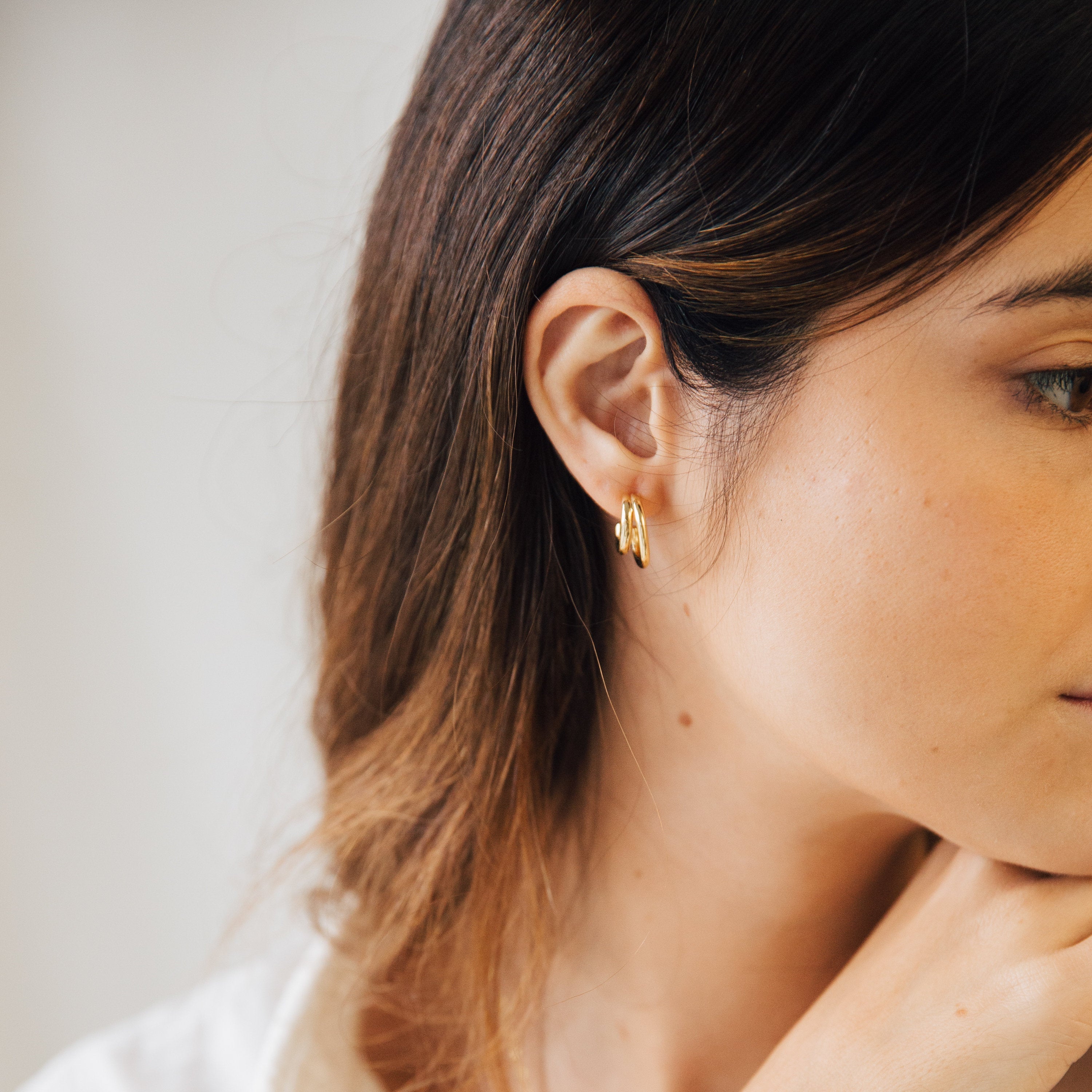 Close-up of a woman with long brown hair wearing Zoey Duo Hoops, resting her chin on her hand and showcasing a subtle stacked ear look.