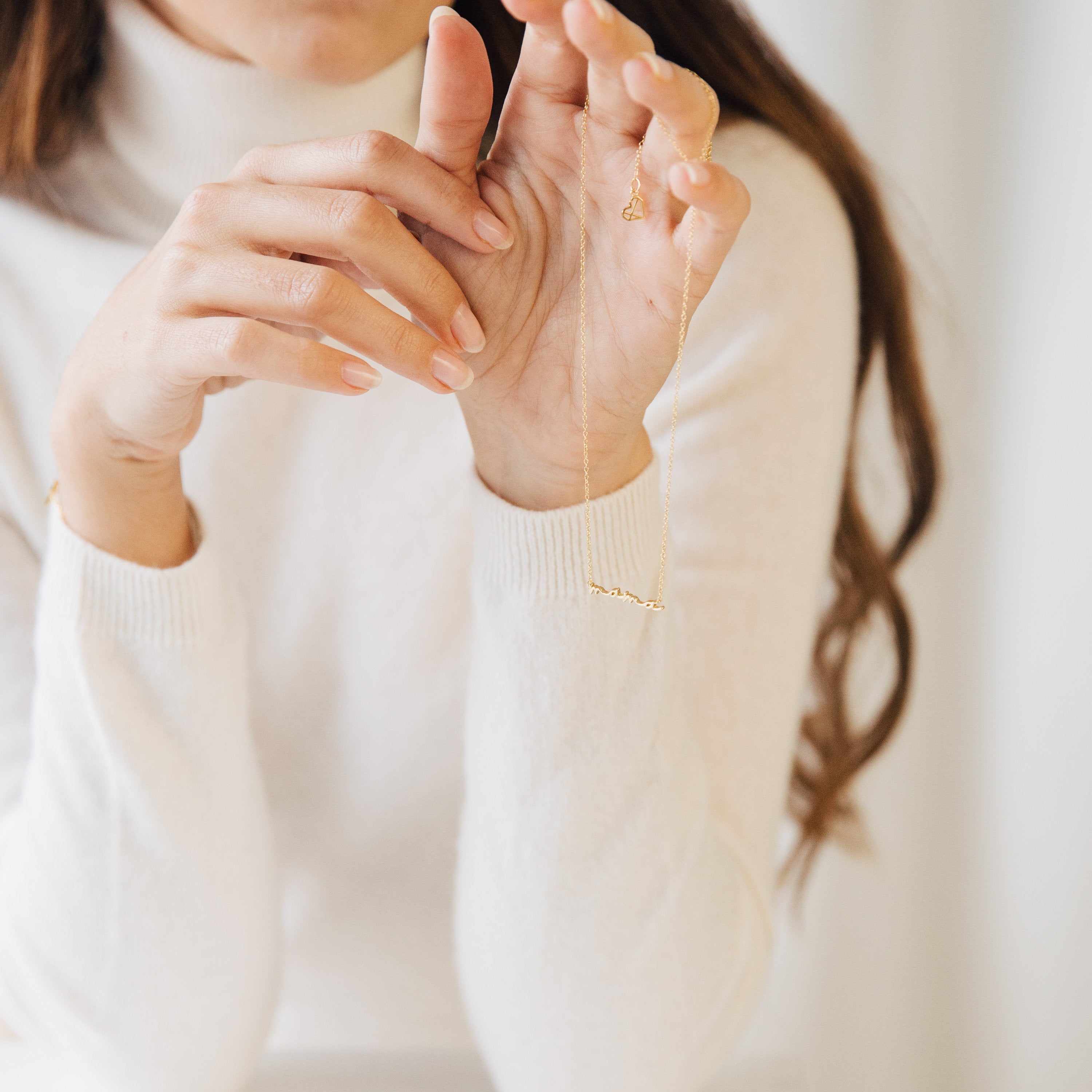 A person in a white turtleneck holds up the Dainty Mama Necklace in 18K Gold, designed in a delicate script, with only their hands and part of their face visible.