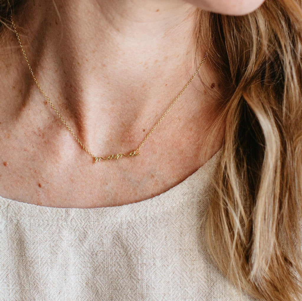 Close-up of a person wearing the Dainty Mama Necklace in 18K Gold, featuring a delicate mama script. The wearer, with light skin and long light brown hair, is dressed in a beige top. This necklace makes a perfect gift.