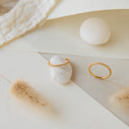 The Dove Eternity Ring and a petite gold ring are displayed on white stones and textured beige paper, accented with dried grasses.