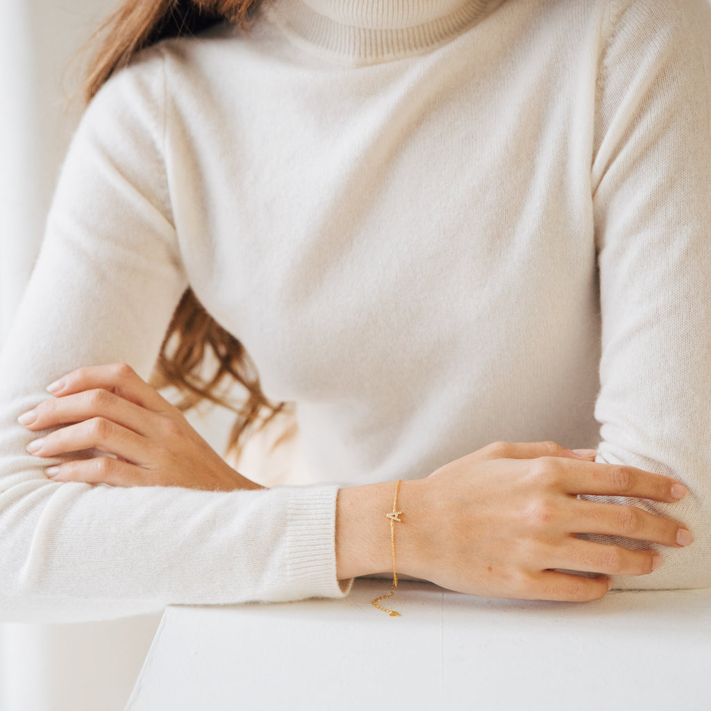 A woman in a cream sweater rests her arms on a table, showcasing the Pave Initial Bracelet on her wrist.