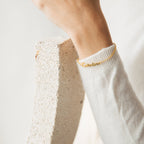 The Ladybirds Name Curb Bracelet, featuring the name Caroline, is shown worn on a wrist with a white sweater against textured stone, highlighting its contemporary curb chain style.