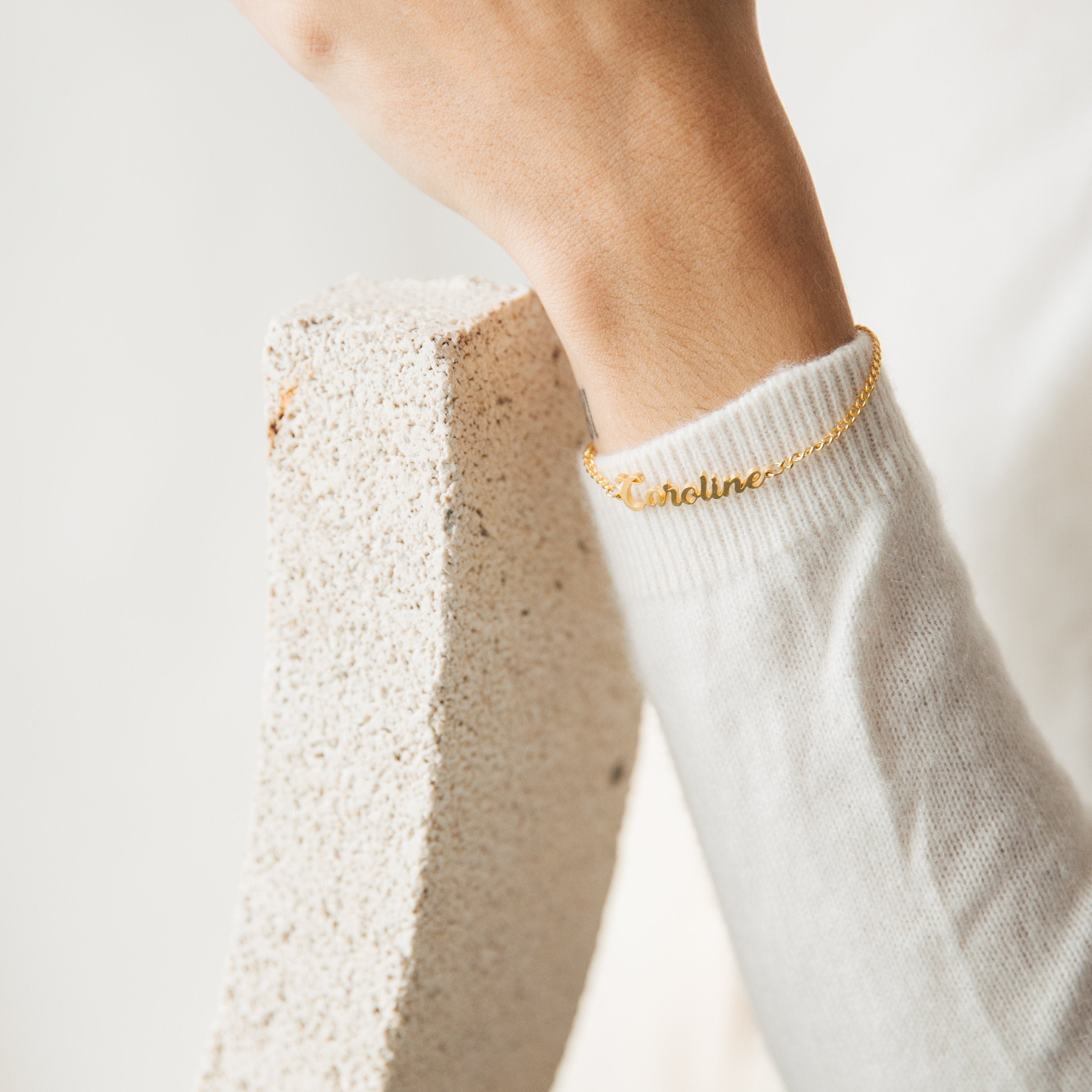 The Ladybirds Name Curb Bracelet, featuring the name Caroline, is shown worn on a wrist with a white sweater against textured stone, highlighting its contemporary curb chain style.