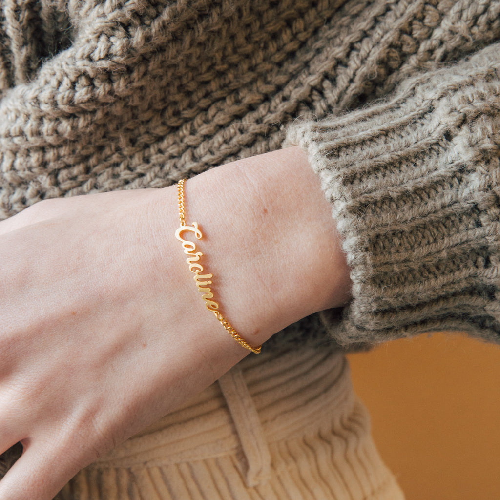 The Ladybirds Name Curb Bracelet with "Caroline" shines on a wrist, styled with a beige knit sweater—an elegant, modern bracelet perfect for any occasion.