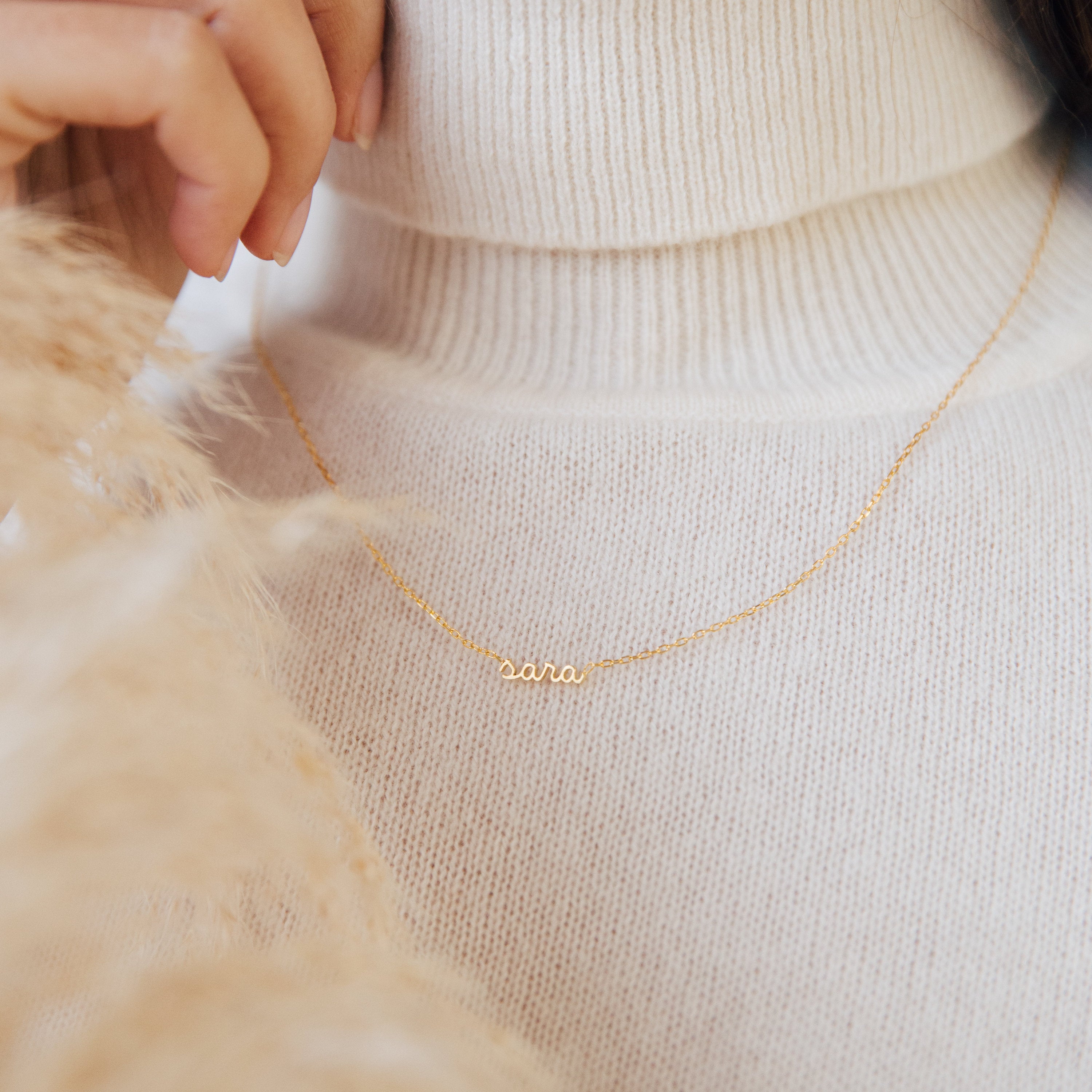 The Petite Tiny Name Necklace with a "sara" pendant, styled over a white turtleneck sweater—an elegant personalized piece and the perfect gift for mom.