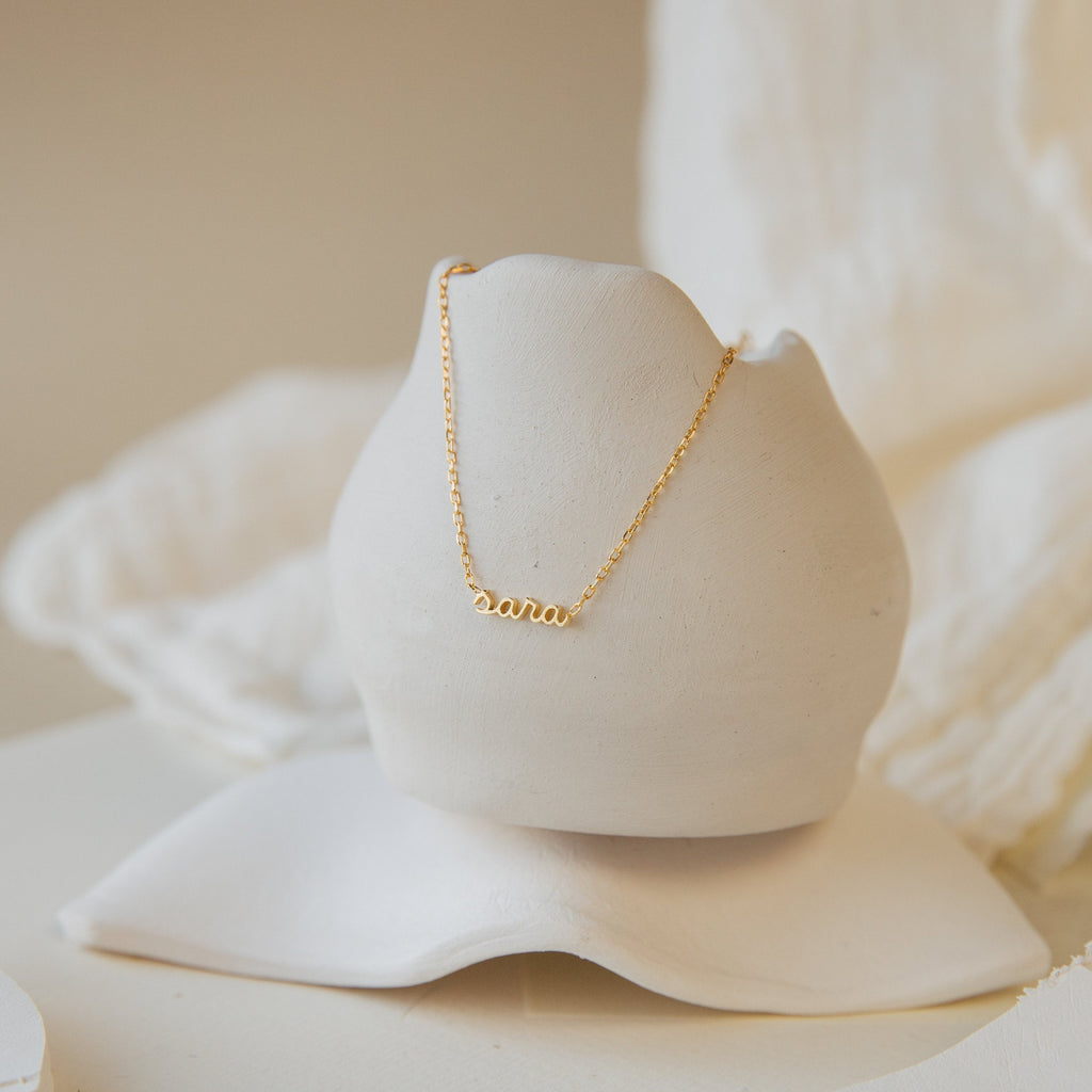 Petite Tiny Name Necklace featuring “ana” displayed on a white ceramic stand against a soft, neutral background—an elegant personalized necklace and perfect gift for mom.