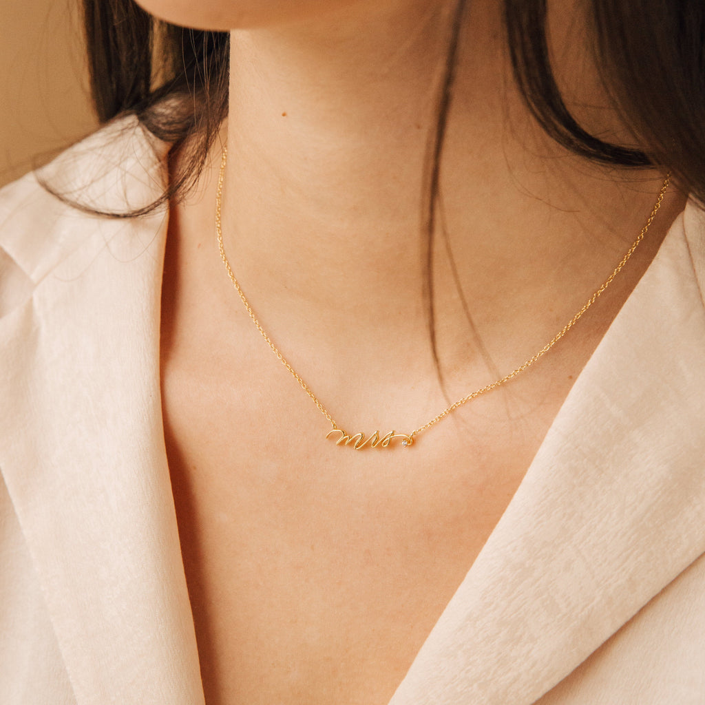 A woman wears the Mrs. Necklace, a 925 sterling silver piece with a delicate script pendant, paired with a light v-neck blouse—an ideal wedding gift.