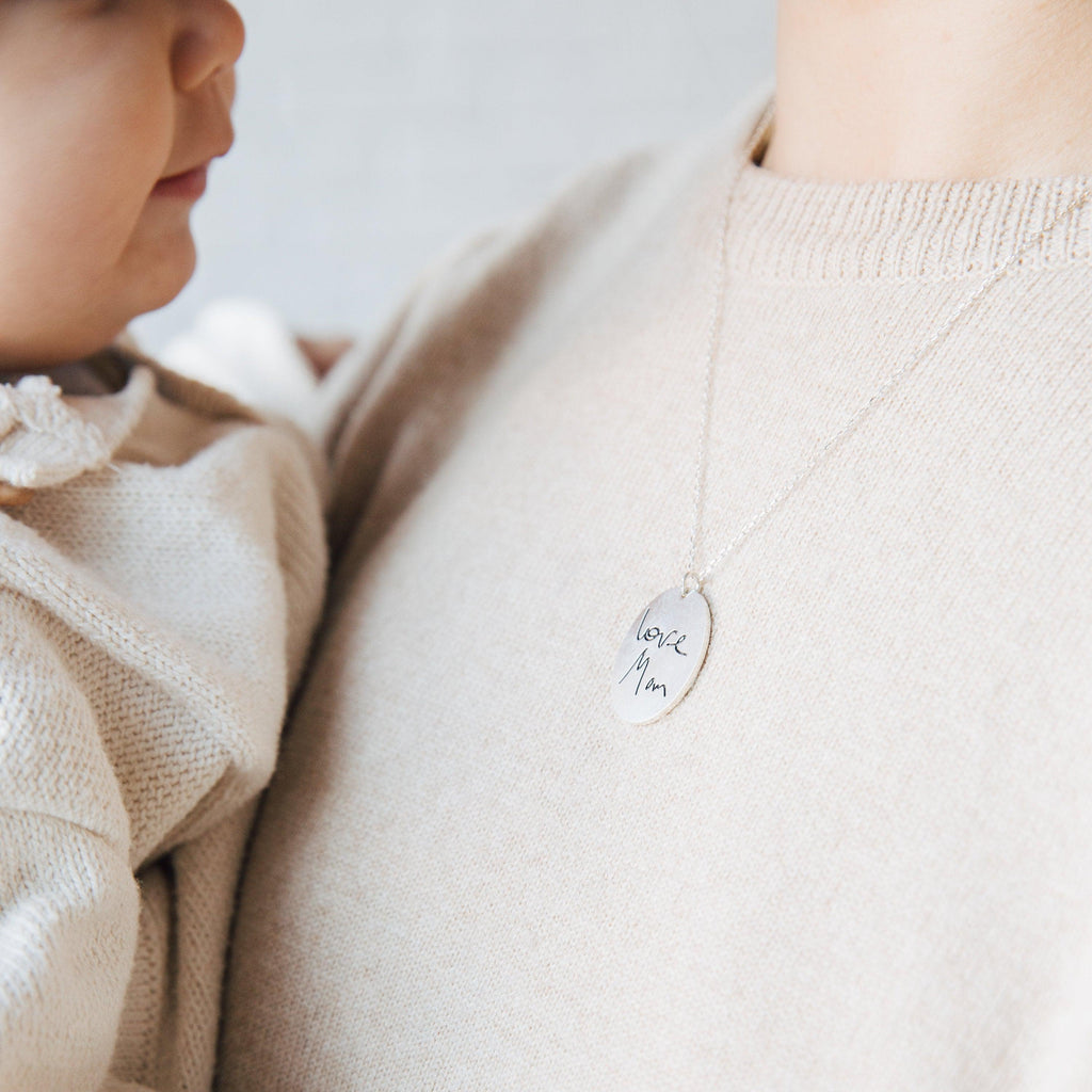 A woman holding a baby wears the Oval Fingerprint Necklace, featuring a personalized fingerprint etching and engraved with "love, Mom" on the pendant.