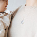 A woman holding a baby wears the Oval Fingerprint Necklace, featuring a personalized fingerprint etching and engraved with "love, Mom" on the pendant.