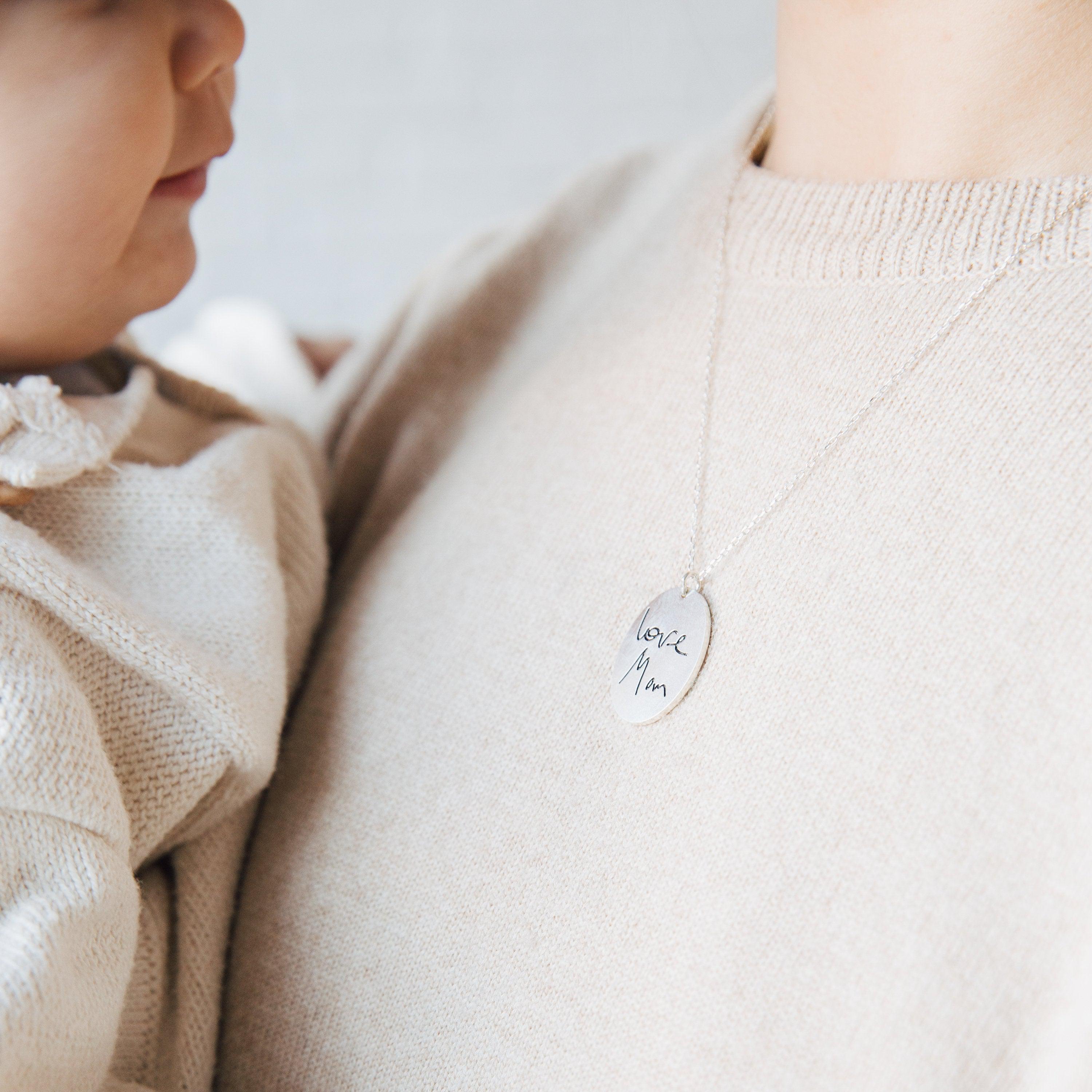 A woman holding a baby wears the Oval Fingerprint Necklace, featuring a personalized fingerprint etching and engraved with "love, Mom" on the pendant.