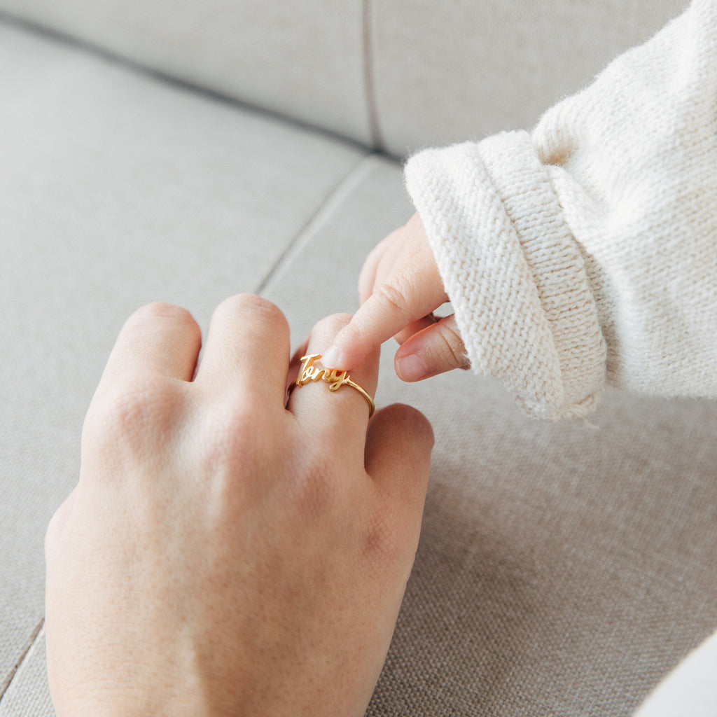 A baby’s hand touches an adult’s wearing the Grace Script Name Ring in Rose Gold, inscribed with “love,” on a light fabric surface.