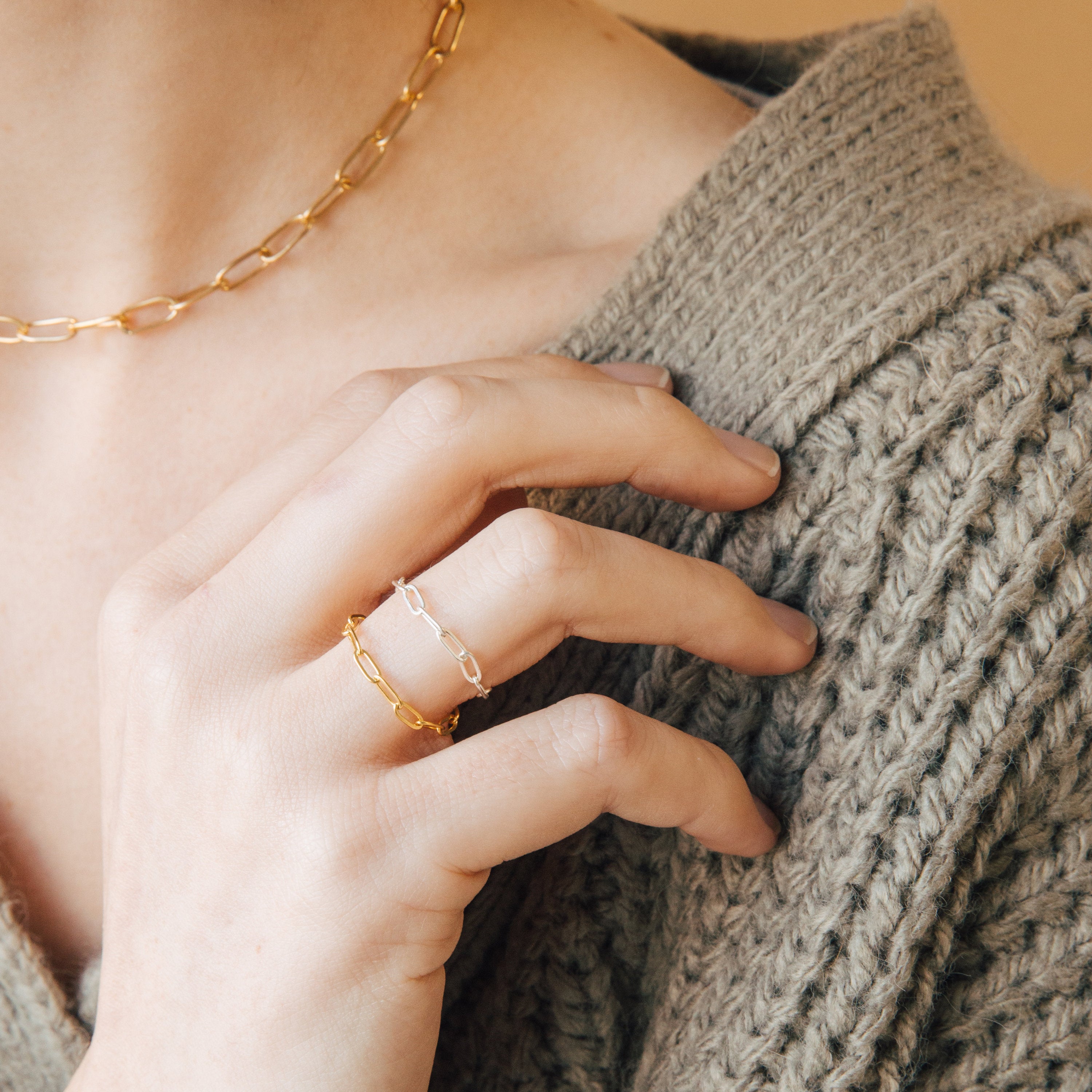 A person wearing a Paperclip Chain Ring and a gold chain necklace, paired with a chunky knitted sweater in beige tones.