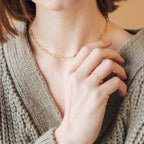 A woman, dressed in a knitted beige sweater, wears stacking rings including the Paperclip Chain Ring and a gold chain necklace.