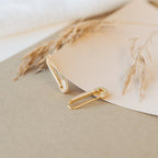 The Luxx Earrings, inspired by gold safety pins, rest on beige paper with dried grass in the background.