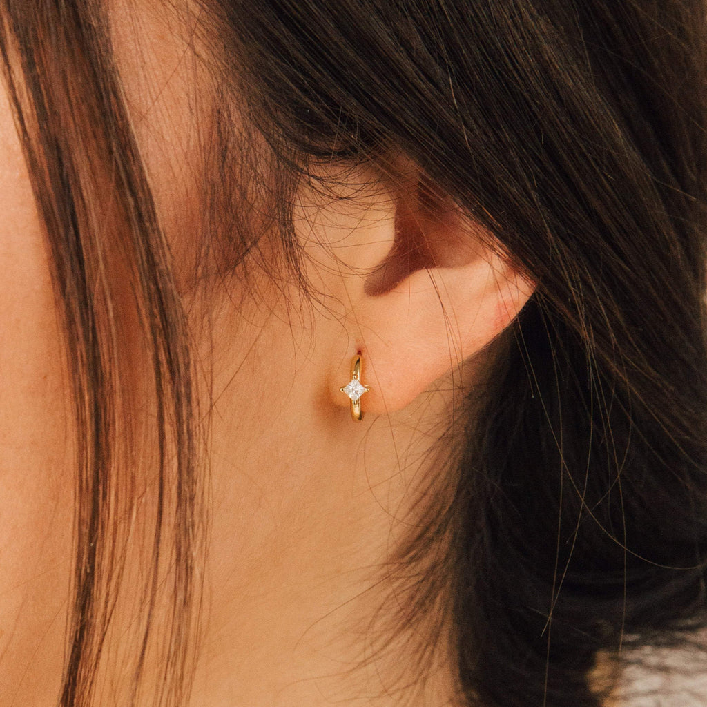 Close-up of a woman's ear adorned with Athena Huggies—gold hoop earrings set with a single diamond-shaped stone.