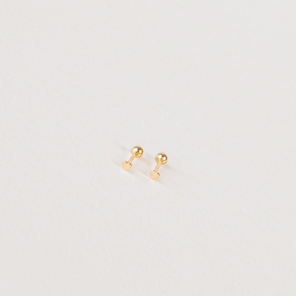 The Tiny Heart Studs in 18K Gold are a perfect gift, featuring delicate heart-shaped gold studs on a white surface—ideal for everyday wear.