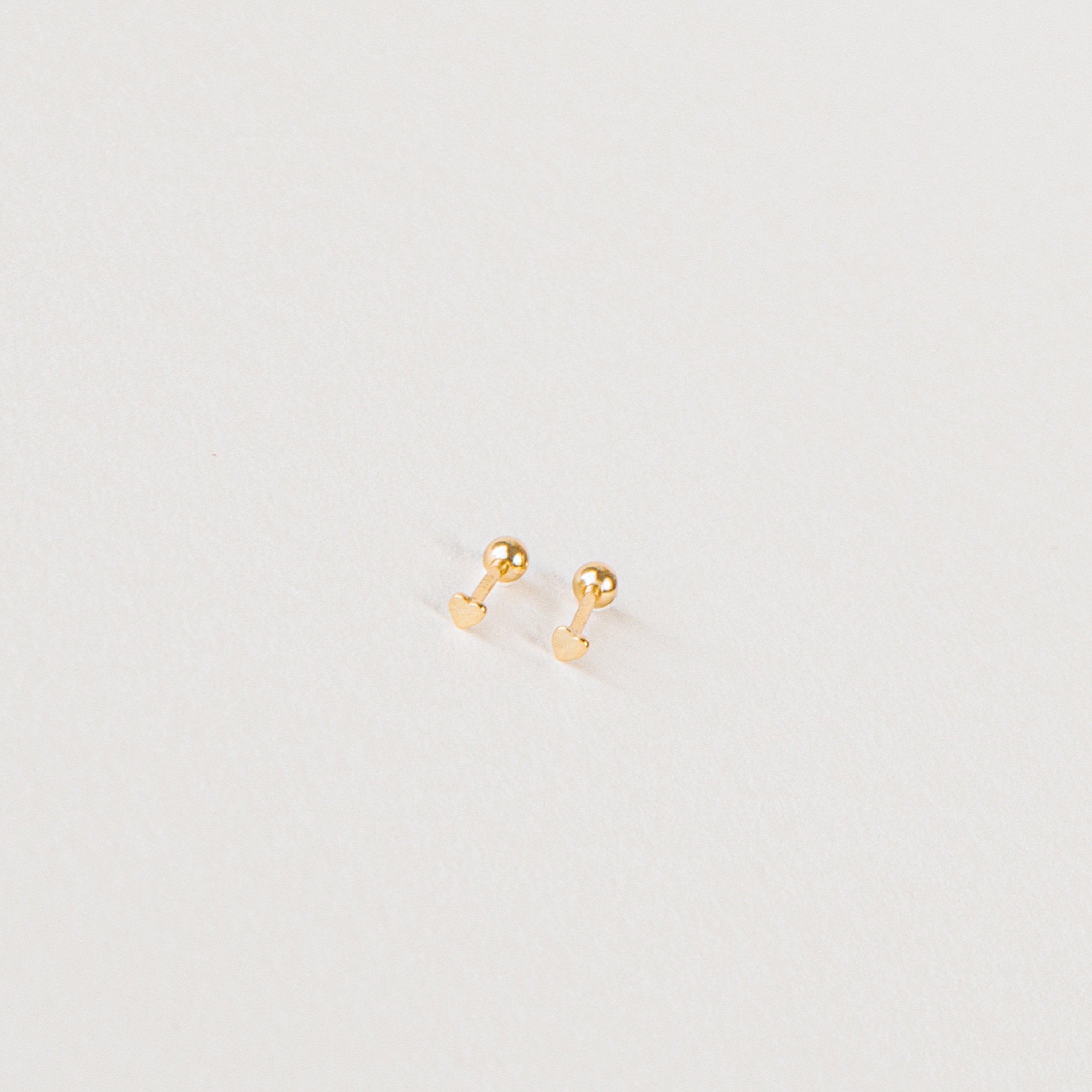 The Tiny Heart Studs in 18K Gold are a perfect gift, featuring delicate heart-shaped gold studs on a white surface—ideal for everyday wear.
