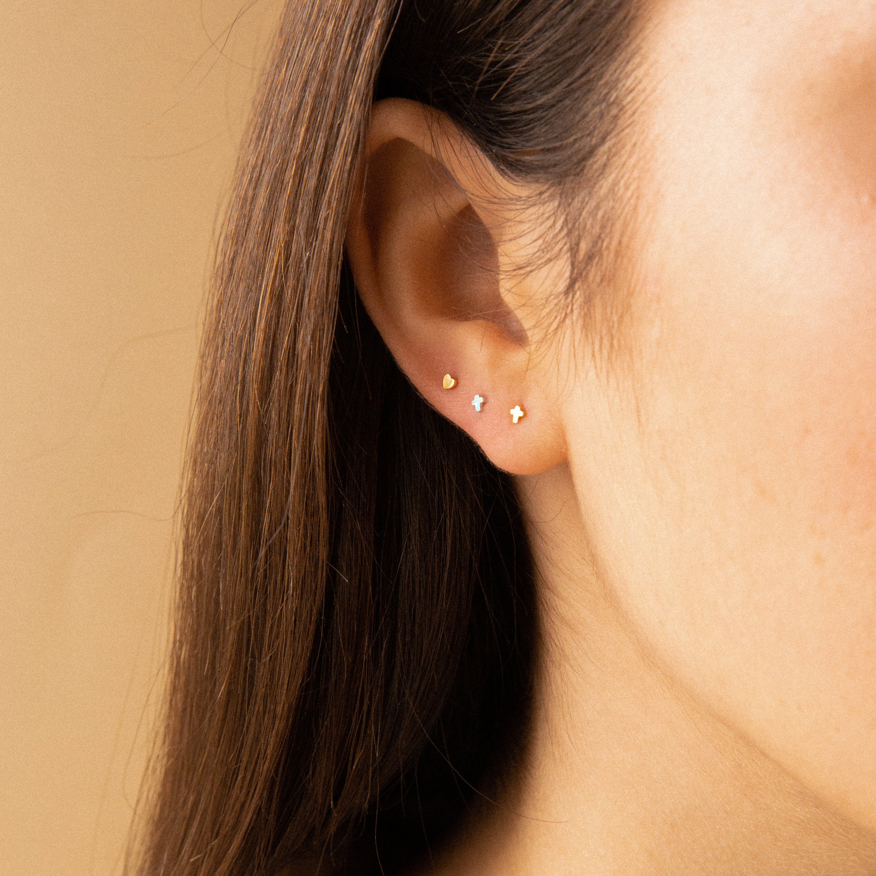Close-up of an ear with three small gold studs: Tiny Heart Studs in 18K Gold, a round stud, and a star. Set against a plain beige background—ideal as a thoughtful gift or chic daily accessory.