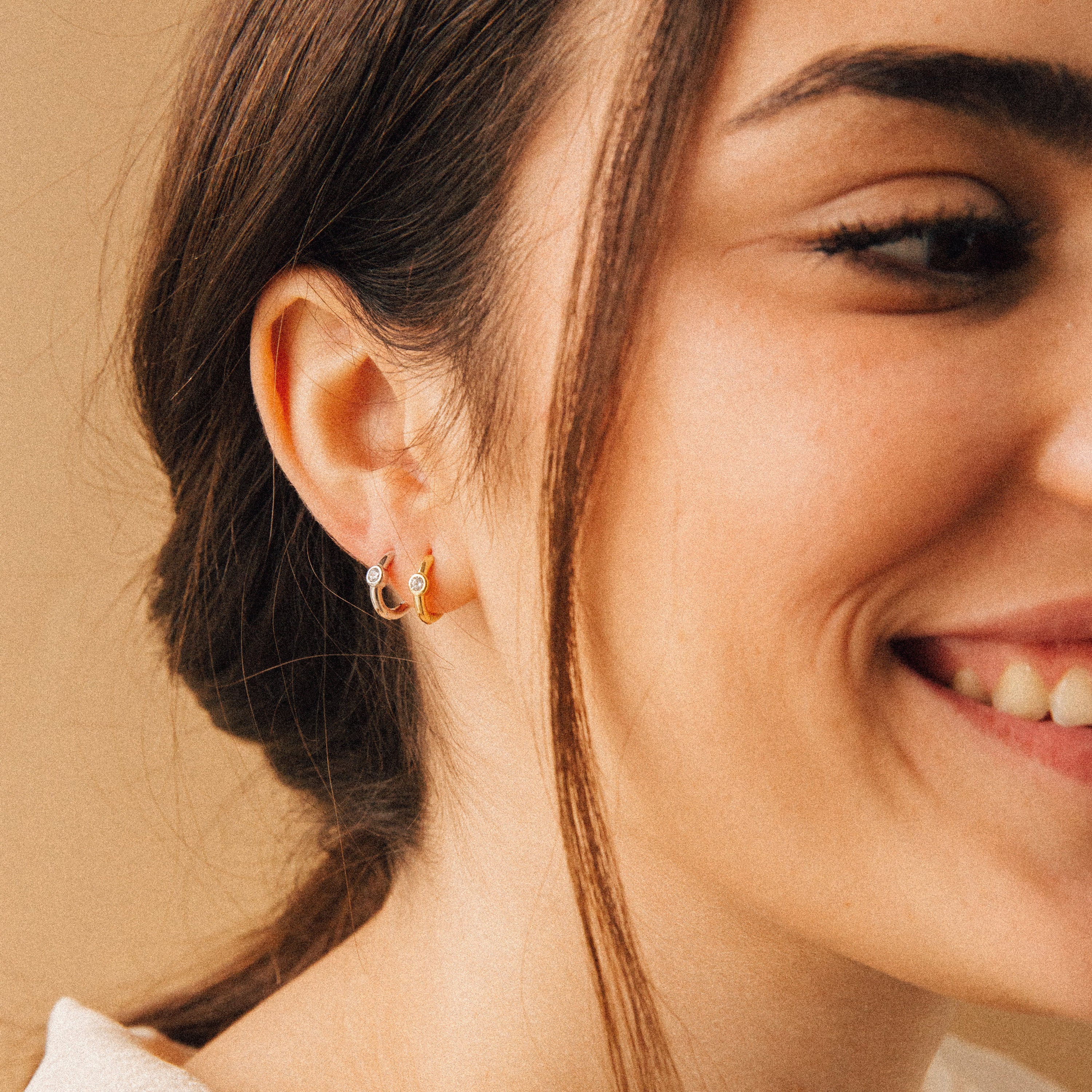 A woman with brown hair smiles, wearing Aela Huggies—elegant small hoop earrings that glint on her ear—set against a beige background.